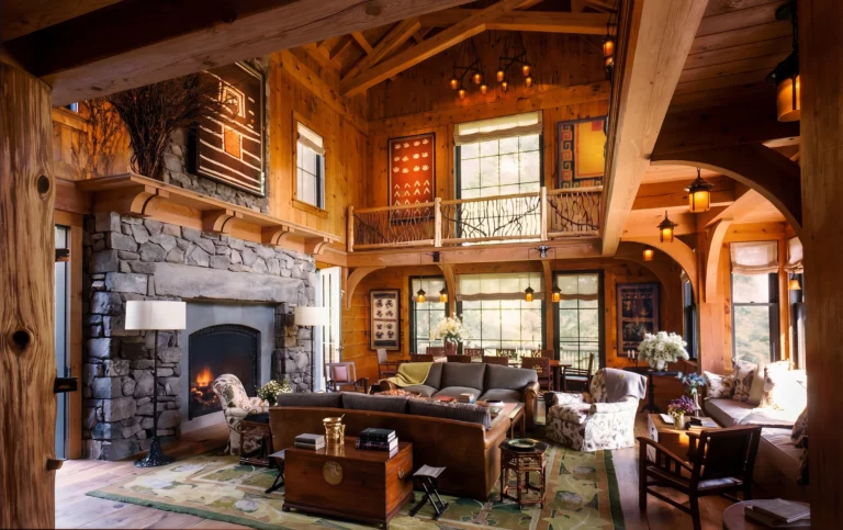 Double-height great room with exposed timber trusses, a large fieldstone fireplace, and lounge seating beneath warm lantern lighting.