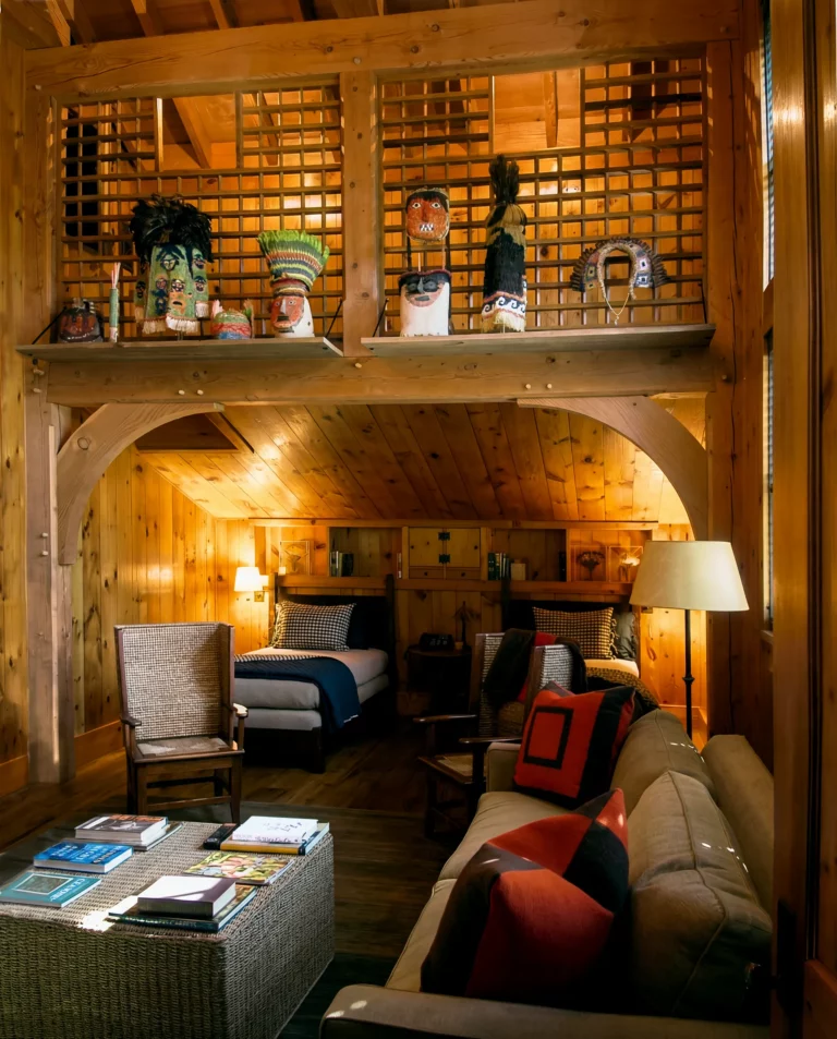 A cozy wooden cabin interior with a loft. The ground floor has a sofa with colorful pillows, wicker chairs, a coffee table with magazines, and a lamp. The loft railing displays decorative masks and crafts—bringing rustic charm to interiors far from Manhattan.