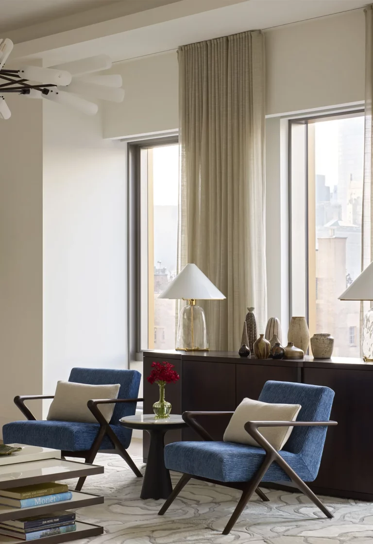 Living room seating vignette in a Snøhetta Upper West Side apartment with blue lounge chairs, a small round table, and soft daylight framed by tall curtains.